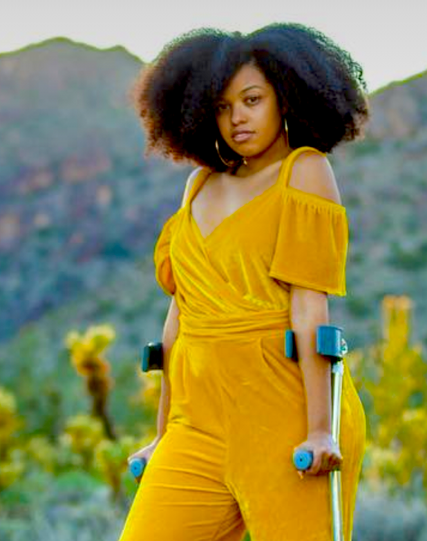 A brown woman with thick, frizzy hair standing on forearm crutches.