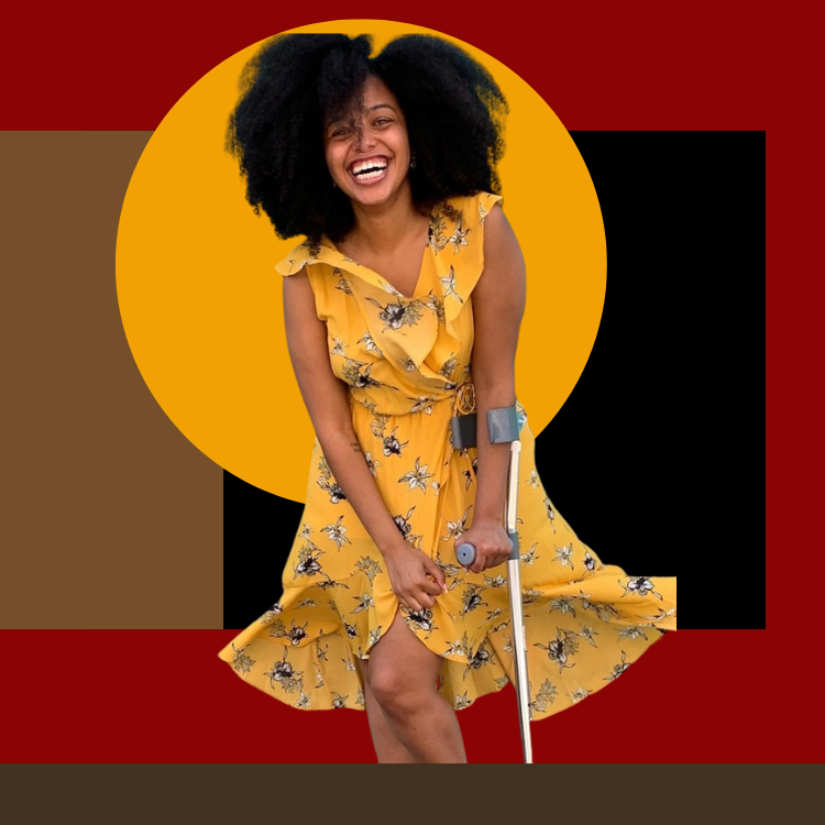 A laughing brown girl using a forearm crutch tries to stop her dress from blowing in the wind with her free hand.