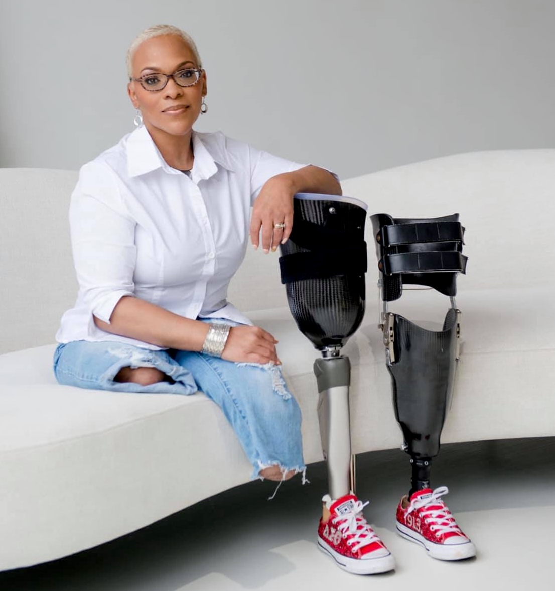 A black female bilateral leg amputee with light skin and short hair is sitting on a white couch. Two prosthetic legs are standing in front of her.