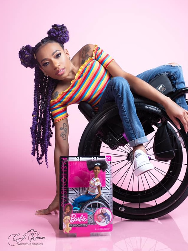 A young black girl leaning back in a wheelchair with a package for a black Barbie doll who uses a wheelchair in front of her.