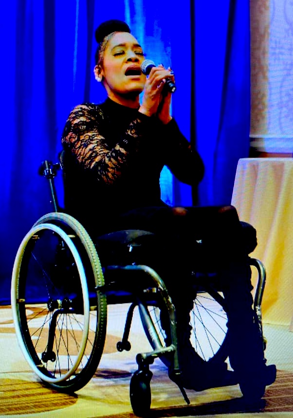 Black woman in a wheelchair holding a microphone near her mouth.